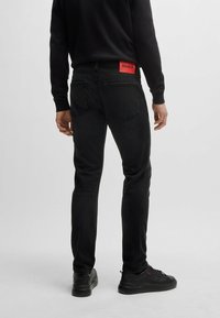 Black denim jeans featured with a red branded tag, slim fit design, five-pocket style, and paired with black sneakers.