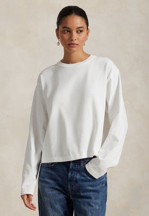 Młoda kobieta o ciemnych włosach, ubrana w białą koszulę z długim rękawem i niebieskie dżinsy, stojąca z jedną ręką w kieszeni na neutralnym tle.