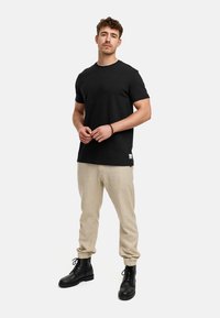 Camiseta negra de manga corta combinada con pantalones beige claros. El modelo lleva botas negras de cordones, mostrando un estilo casual.