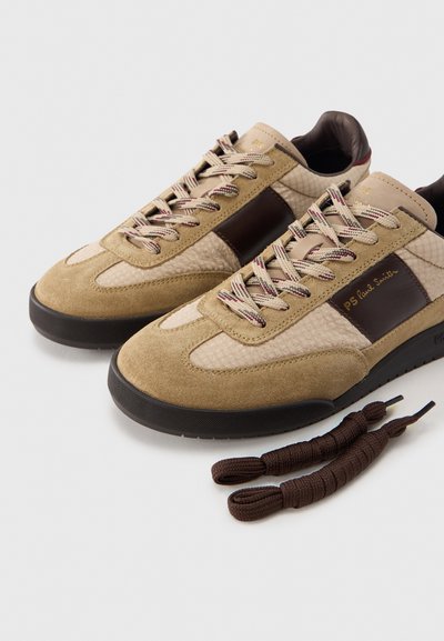 Τα μπεζ και καφέ σουέτ αθλητικά παπούτσια διαθέτουν λεπτομέρειες από δίχτυ, πλεγμένα κορδόνια και μαύρη  λαστιχένια σόλα. Περιλαμβάνουν επιπλέον σκουρόχρωμα καφέ κορδόνια.