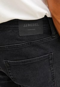 Jeans in denim neri con superficie texturizzata, tasche posteriori e una patch rettangolare in pelle nera con la scritta "JJ REBEL" in rilievo.