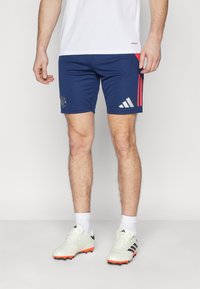 Adidas tmavomodré futbalové šortky s červenými bočnými pruhmi, detailom loga, elastickou tkaninou a priliehavým strihom, v kombinácii s bielymi futbalovými kopačkami.