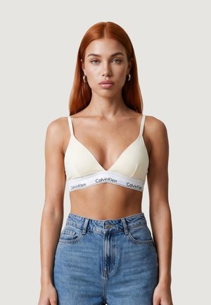 Mladá žena s dlouhými červenými vlasy, která má na sobě bílý bralette Calvin Klein a modré džíny s vysokým pasem, čelí dopředu na jednobarevném pozadí.