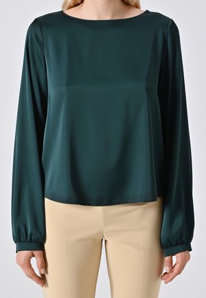Femme portant un chemisier vert foncé à manches longues avec un pantalon beige, debout devant un fond clair uni.