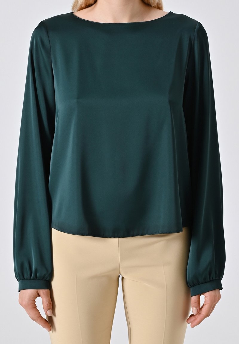 Femme portant un chemisier vert foncé à manches longues avec un pantalon beige, debout devant un fond clair uni.