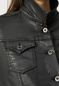 Schwarze Denimjacke mit hohem Kragen, Druckknöpfen und zwei vorderen Taschen mit Klappenverschluss. Der Stoff hat einen glänzenden Finish.