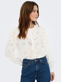 Blouse blanche dotée de découpes florales et de détails en dentelle, avec un col rond et des manches longues froncées, associée à un jean taille haute.