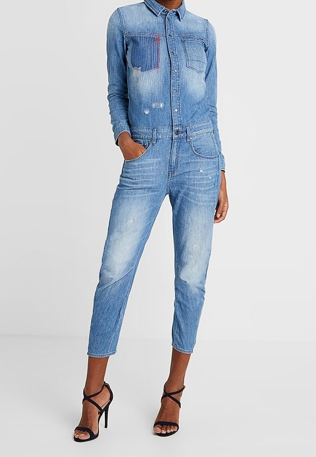 Femme portant une combinaison en denim bleu délavé avec des détails en patch et des sandales noires à talons hauts et à brides, debout avec une main dans la poche.