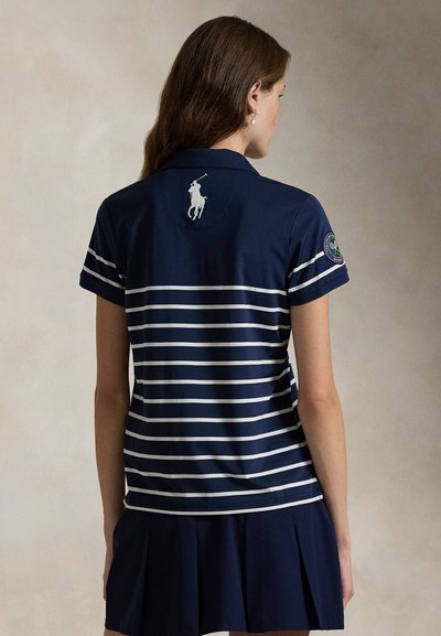 Donna che indossa una polo a maniche corte blu navy con strisce orizzontali bianche, logo del giocatore di polo bianco sulla schiena e toppa di Wimbledon sulla manica.