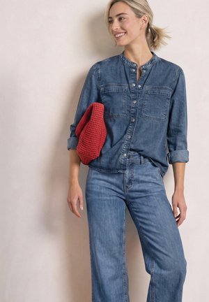Smilende kvinde med blond hår i lav hestehale iført denimskjorte og jeans, der holder en foldet rød strikket taske under armen.