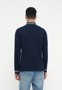 Navy long-sleeve polo shirt featuring a two-tone collar with green and white stripes. Paired with light blue denim jeans.