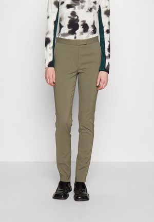 Persona che indossa pantaloni slim fit verde oliva, scarpe nere lucide e una maglia bianca a maniche lunghe con motivi astratti neri e verde scuro.