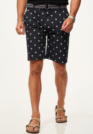 Mann trägt schwarze Shorts mit weißem Palmenmuster, gestreiften Gürtel, schwarzes Hemd, Uhr und beige Sandalen, steht vor weißem Hintergrund.