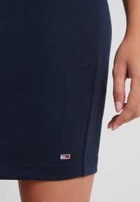 Sukně z modrého bavlněného žerzeje, přiléhavý design, hladká textura, s malým výšivkovým logem v červené, bílé a modré barvě na lemu.