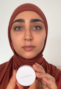 Okrą container kosmetyczny w kolorze białym z etykietą "rms beauty" trzymany przez osobę w rudym hijabie. Gładka tekstura, minimalistyczny design.