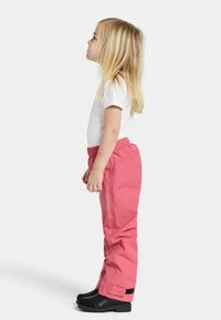 Nuori tyttö seisoo sivuttain valkoisessa lyhythihaisessa paidassa, vaaleanpunaisissa housuissa ja mustissa saappaissa tasaisella taustalla.