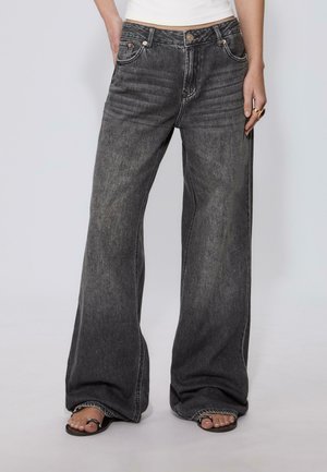 Persona che indossa jeans neri a gamba larga sbiaditi e sandali neri aperti, in piedi con una mano in tasca su uno sfondo semplice.