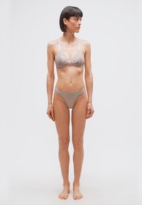 Γυναίκα όρθια ξυπόλυτη με σουτιέν ταφτέ με δαντέλα και ταίριασμα σλιπ, κοιτάζοντας προς τα δεξιά της, μπροστά σε απλό λευκό φόντο.