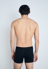 Caleçons boxeurs noirs en tissu lisse, avec une ceinture et une coupe ajustée, présentés sur le dos nu d'un modèle contre un fond clair.
