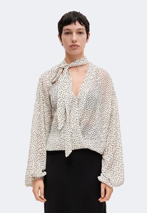 Femme aux cheveux courts et foncés porte un chemisier blanc à pois avec un col noué et des manches longues, assorti à une jupe noire.