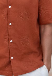 Rust-colored short-sleeve shirt with embroidered sun motifs, featuring white buttons and a textured fabric. Accents include subtle stitching details.
