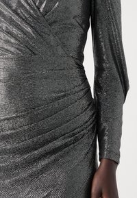 Robe argentée brillante à manches longues, présentant un design froncé et un tissu texturé. La surface reflète la lumière, renforçant l'aspect général.