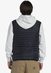 Padded black vest with quilted design, worn over a grey hoodie. Features a high collar and no sleeves, providing insulation without bulk.