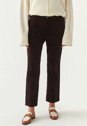 Bruine corduroy broek met een aansluitend ontwerp, hoge taille en voorzak. Gedirigeerd met een crèmekleurige trui en bruine instappers met gouden accenten.