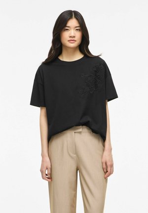 Kvinde med langt mørkt hår iført en sort t-shirt med blomsterbroderi og beige bukser, stående foran en hvid baggrund.