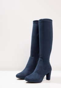 Granatowe, zamszowe kozaki do kolan o gładkiej fakturze, zaokrąglonym nosku i średniej wysokości obcasie; posiadają zamek błyskawiczny z boku do zapinania.