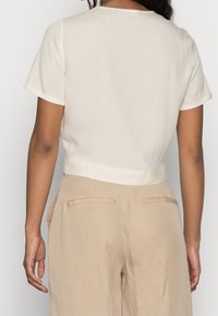 Mujer con blusa de manga corta color crema y pantalones beiges de talle alto, vista desde atrás contra un fondo blanco liso.