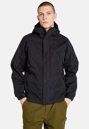 Giacca impermeabile nera con cappuccio, dotata di due tasche frontali, chiusura a scattino e polsini regolabili. Indossata sopra pantaloni verde oliva.