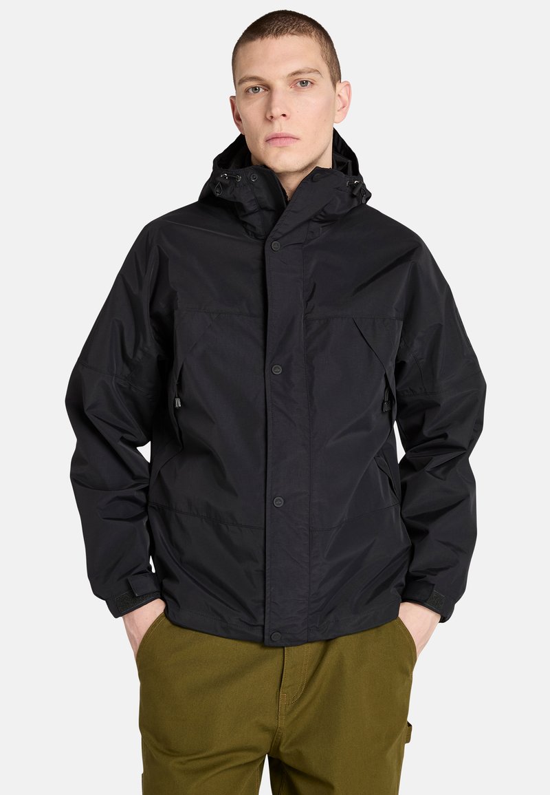 Mustat vedenpitävät takki, jossa on huppu, kaksi etutaskua, neppari kiinnitys ja säädettävät hihansuut. Päällä oli oliivinvihreät housut.