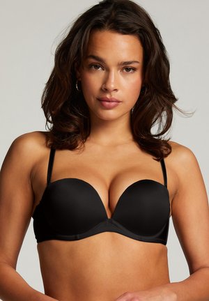 Frau mit welligem dunklem Haar, die einen schwarzen gepolsterten BH mit dünnen Trägern trägt, frontal vor einem schlichten hellen Hintergrund.