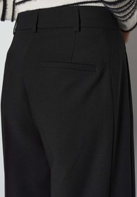 Pantalon noir en tissu lisse, avec une taille slim, des passants de ceinture et une poche arrière unique. Design épuré sans motifs visibles.