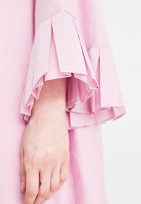 Ροζ φόρεμα με πτυχές και φουσκωτά μανίκια. Το ύφασμα φαίνεται λείο με ελαφριά γυαλάδα. Ένα χέρι με ένα μικρό τατουάζ είναι μερικώς ορατό.