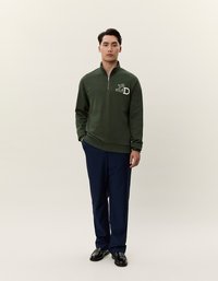 Felpa con zip verde scuro con logo bianco "LD", abbinata a pantaloni blu navy e scarpe nere, su uno sfondo neutro.