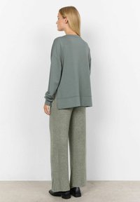 Losse, lange mouwen, salie groene trui met zijsplitten, gecombineerd met wijde, textuur pants in dezelfde kleur en platte zwarte schoenen.