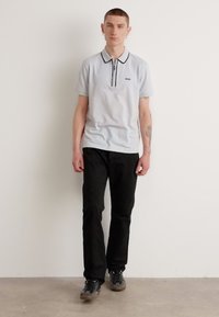 Polo shirt grigio chiaro con rifiniture nere, piccolo logo sul petto, abbinato a pantaloni neri e scarpe da ginnastica grigie, in piedi contro uno sfondo semplice.