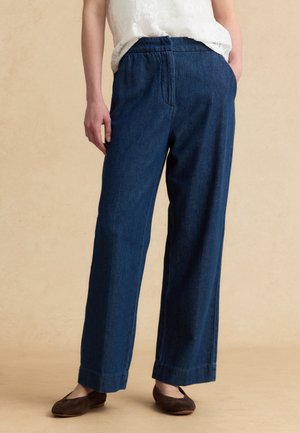 Woman wearing dark blue wide-leg denim pants with elastic waistband and brown flat shoes, standing against beige background.