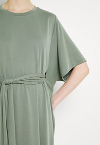 Personne portant une robe ample vert sauge à manches courtes avec une ceinture en tissu nouée à la taille, sur un fond uni.