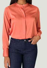 Blouse en satin corail avec un col mandarin et des manches longues, comportant une fermeture à boutons sur le devant et des boutons aux poignets, associée à un jean bleu foncé.