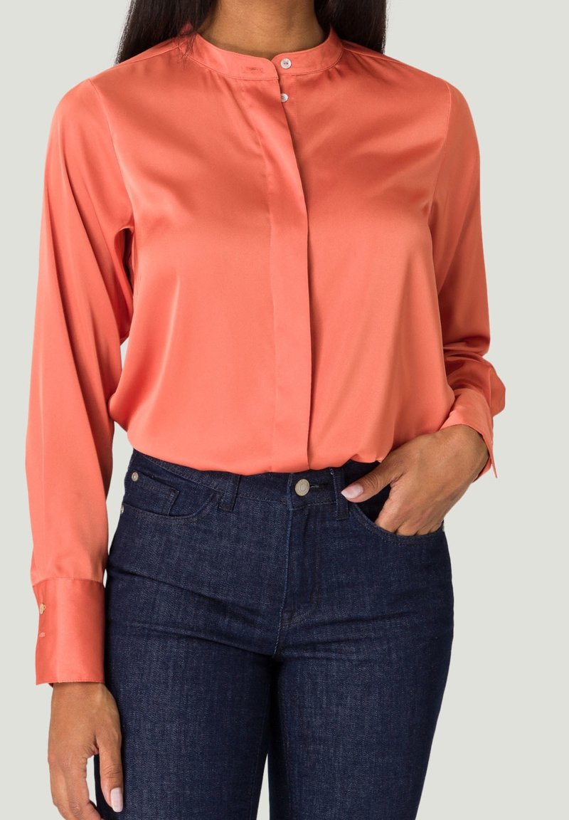 Blouse en satin corail avec un col mandarin et des manches longues, comportant une fermeture à boutons sur le devant et des boutons aux poignets, associée à un jean bleu foncé.