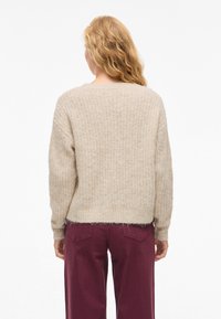 Bež pulover iz pletenine z teksturirano površino, okroglim izrezom in rahlo pristrižnim robom, kombiniran z globoko bordo hlačami.