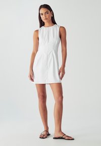 Biele bezrukávové šaty so štruktúrovaným dizajnom, skladmi v oblasti pása a hladkou textúrou; spárované s čiernymi plochými sandálmi.