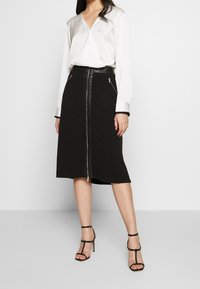 Woman wearing a white long-sleeve blouse, black midi skirt with front zipper and pockets, and black high-heeled sandals standing on a white background.
