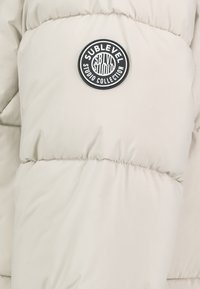 Lichtbeige donsjack met een gequiltde textuur. Beschikt over een ronde zwart-wit logo patch met de tekst "SUBLEVEL" en "STUDIO COLLECTION".
