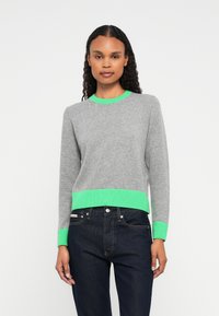 Mulher vestindo um suéter cinza com punhos, decote e bainha verde brilhante, combinado com jeans azul escuro, em pé contra um fundo branco.
