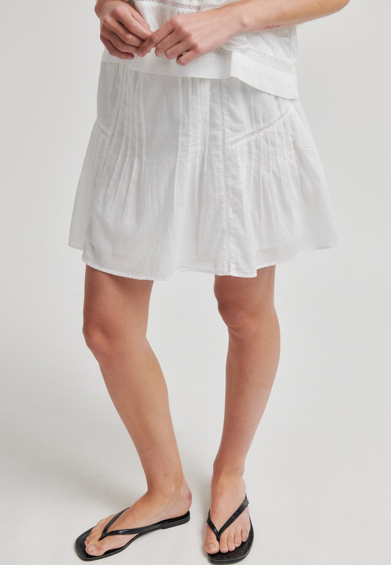 Femme portant une jupe plissée blanche et des tongs noires, debout les mains jointes près de la taille sur un fond uni.