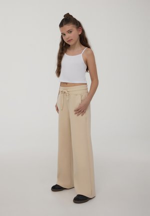 Top corto bianco abbinato a pantaloni larghi beige con vita elastica e lacci, dotati di tasche laterali. Scarpe nere.